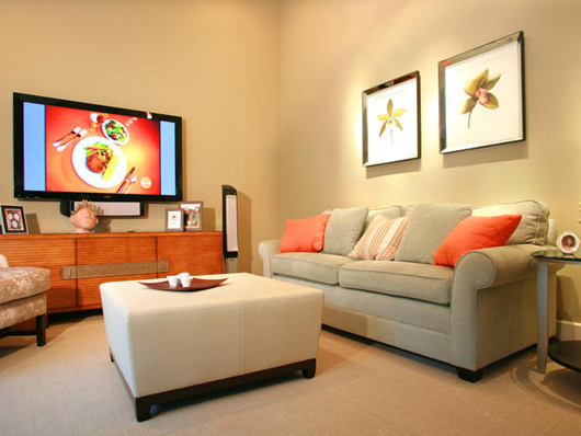 Family room with couch, gray and orange pillows, large flat screen tv mounted on wall, framed art on wall, and carpeted floor.